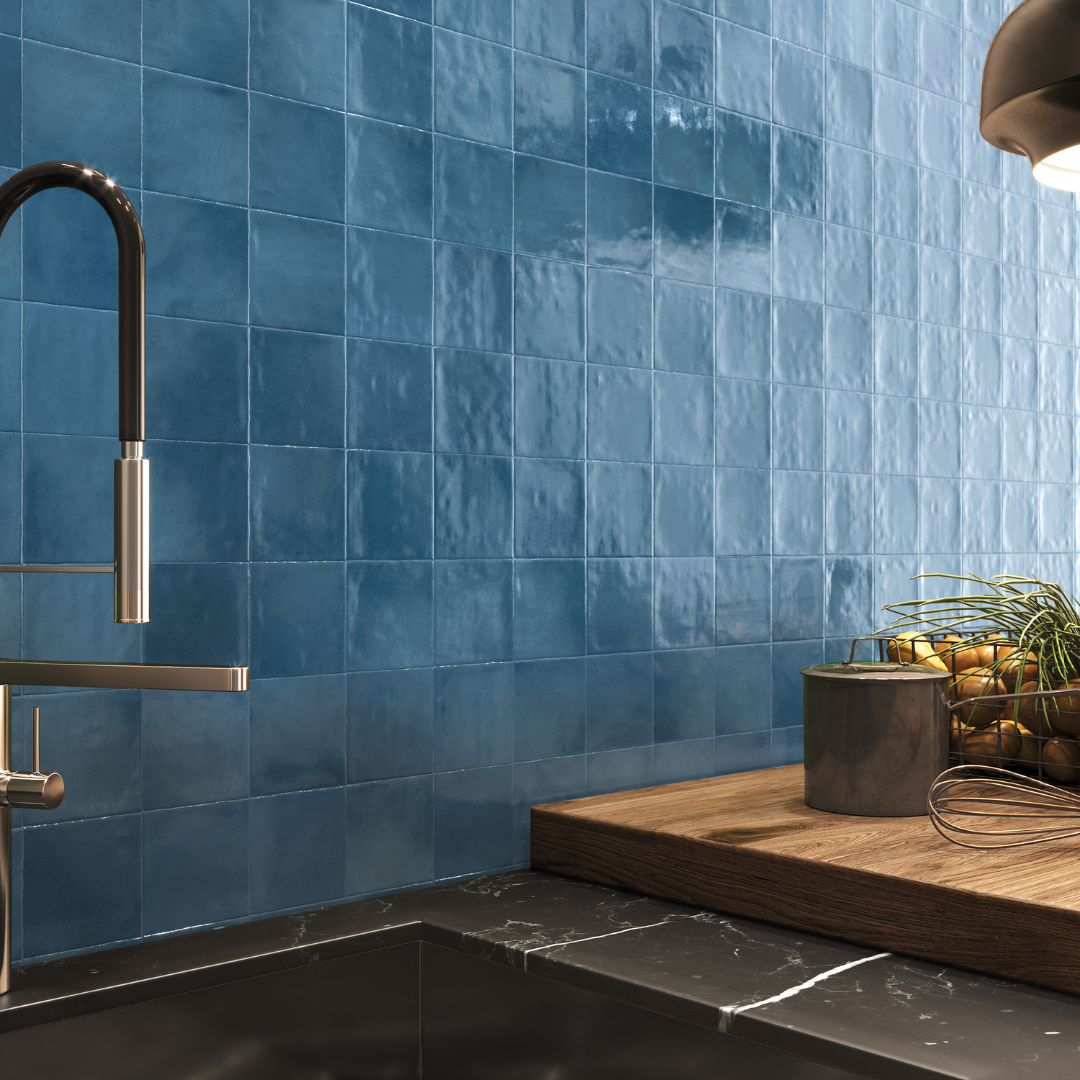 Angled close up of a backsplash tile, modern black kitchen and wood cutting board. The tile on the wall is 4x4 glossy sapphire blue tiles that have textured edges, a slight colour variance and a shiny finish that catches the light. 