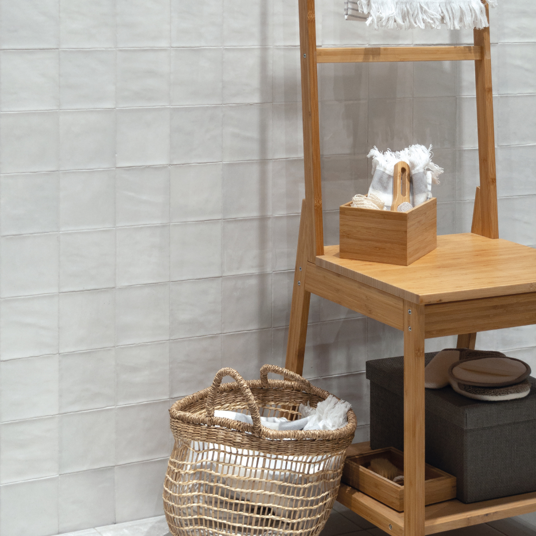 A neutral bathroom wall with a towel wooden leaning towel rack, woven baskets and wood bathroom accessories. The tiled wall is white 4x4 tiles with texture and a glossy finish. 