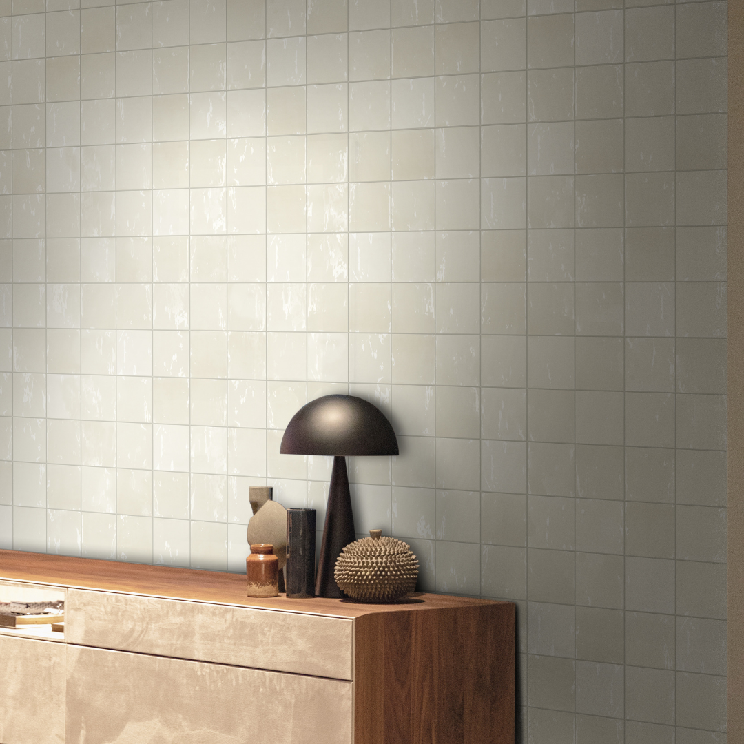 An interior installation of wall tiles with a modern black lamp and wooden credenza. The 5x5 glossy zellige tiles on the wall feature biscuit beige warm shades and a subtle texture. 