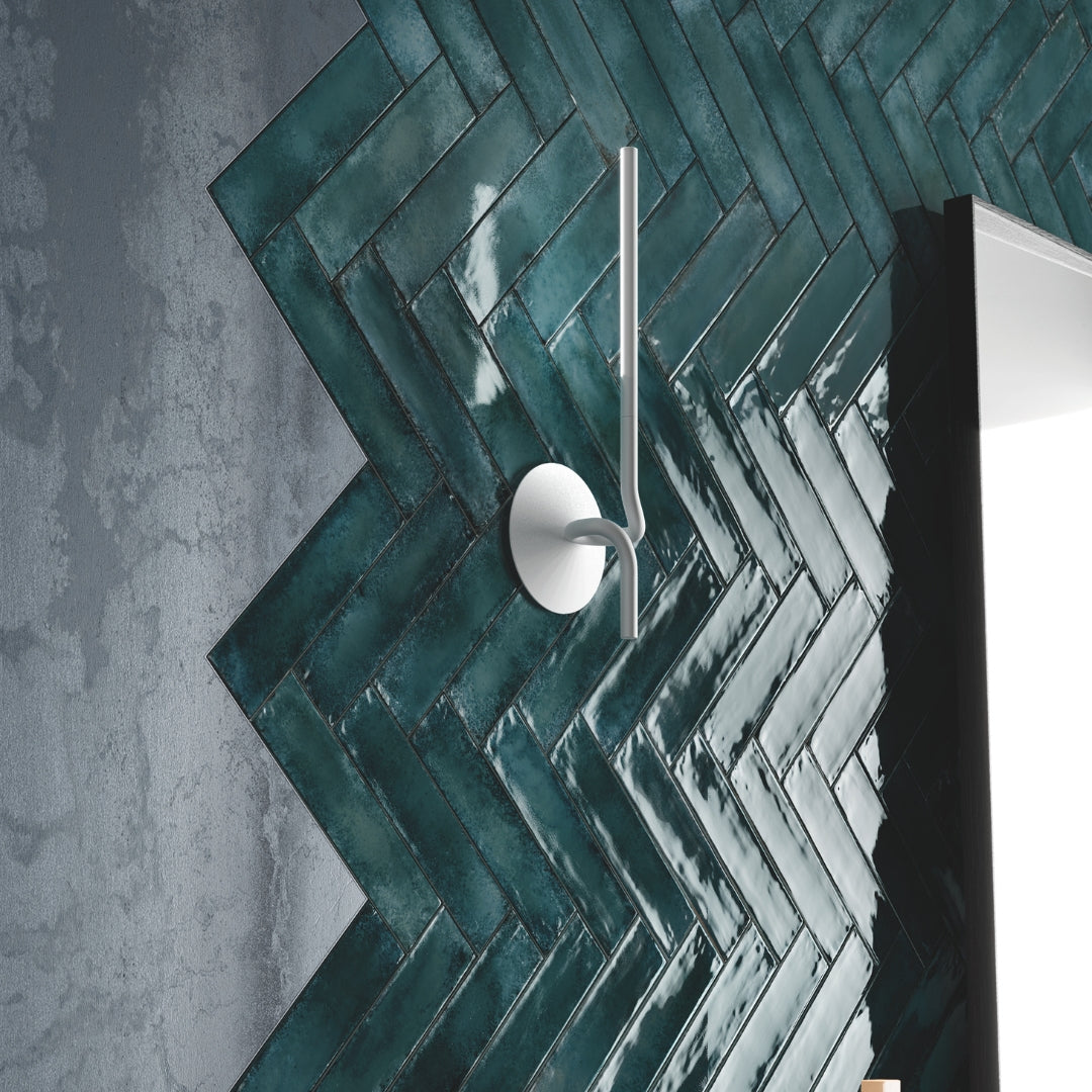 A modern backsplash installation close up. There is a mirror and sconce and grey walls. The back splash tile is a green glossy 2x9 tile laid out in a west to east herringbone pattern