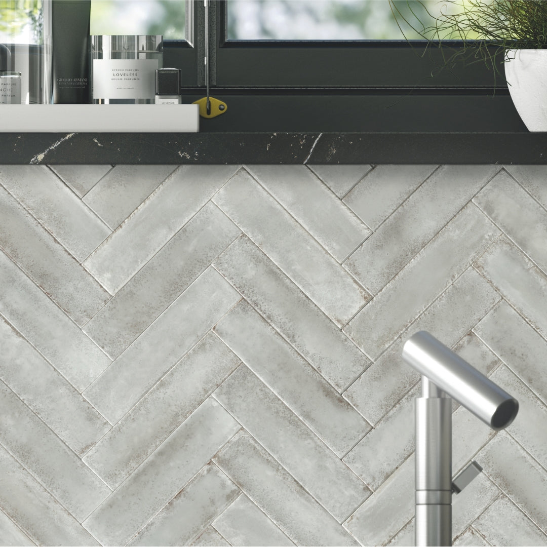 A bathroom backsplash with a black shelf mirror combo and a chrome facet. The tile backsplash is installed in a herringbone pattern, the tiles are grey 2x9 glossy tiles with a rustic charm. 