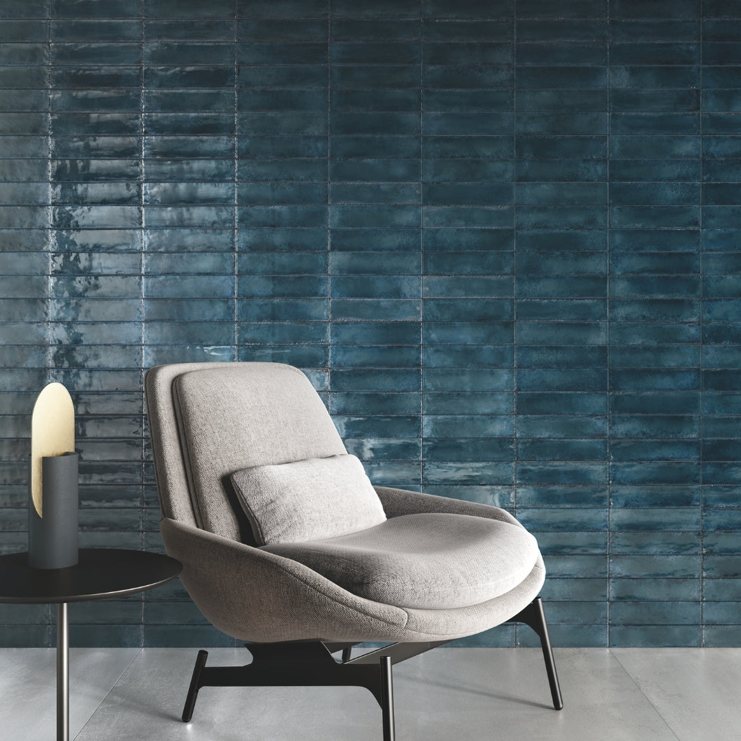 A tranquil seating area with a comfy grey chair with a pretty tile installation behind it. The tiles are 2x9 blue glossy tile with depth and colour variation. The tiles are installed in a modern horizontal stack. 
