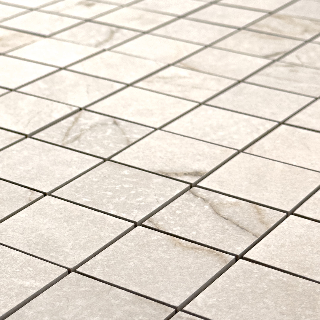 Angled close up view of a 2x2 square sheet mosaic tile that is a cream avorio matte marble look with thin griege veining