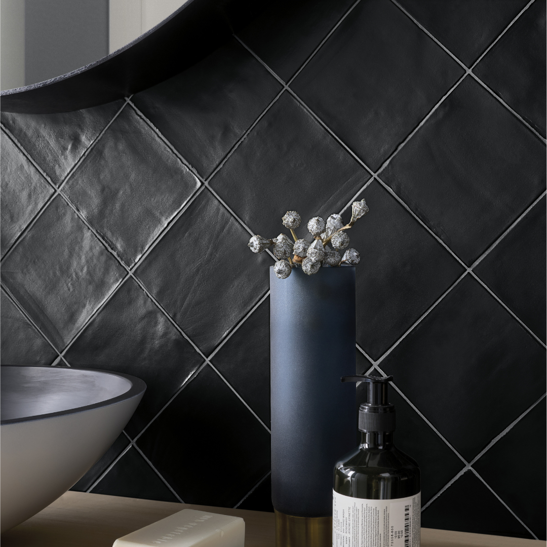 A close up of a bathroom vanity with a matte black tiled backsplash. The tiles are installed in a diamond pattern, the surface is uneven and the edges of tile are tumbled. 