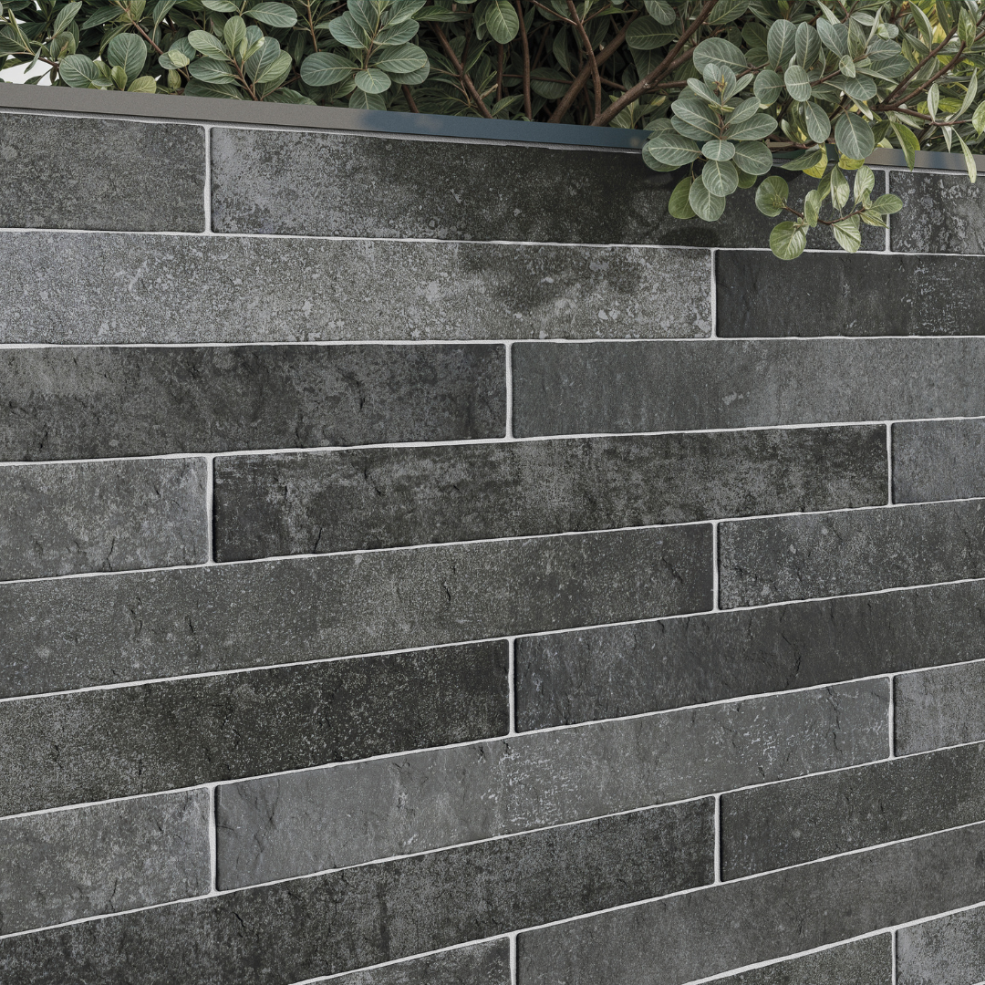 A close up view of an exterior application of tiles on a planter box. The greenery pops against the charcoal black 2x18 brick look tiles that are installed in a traditional horizontal brick pattern. There is colour variation and textured look in the tiles. 