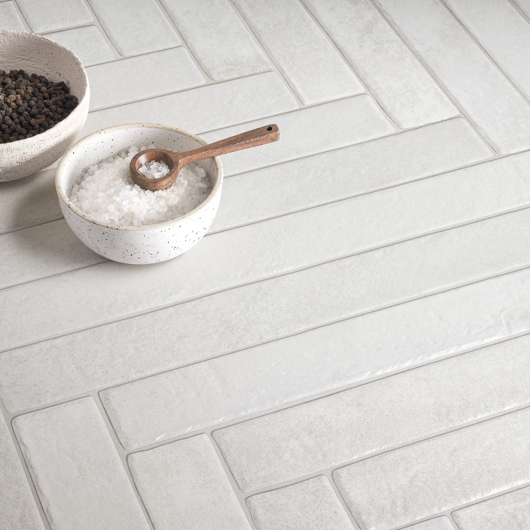 An angled view of a tiled surface, the tiles are white 2x18 and have the appearance of brick stone. They are installed in a herringbone pattern. 