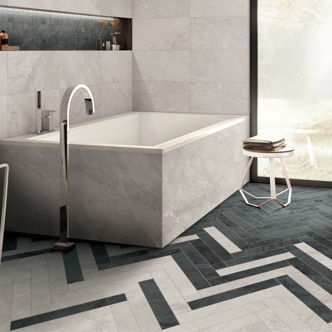A modern bathtub tile installation, the floor of the bathroom showcasing dark grey ad white stone look 4x24 tiles in a herringbone installation. The bathroom walls featuring white stone 12x24 tiles and the niche is tiled with a 2x2 dark grey mosaic backsplash. 