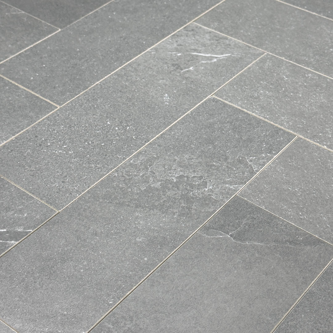 Angled close up of a group of limestone look 4x12 matte tiles in dark grey laid out in a herringbone pattern to show the pattern and finish.