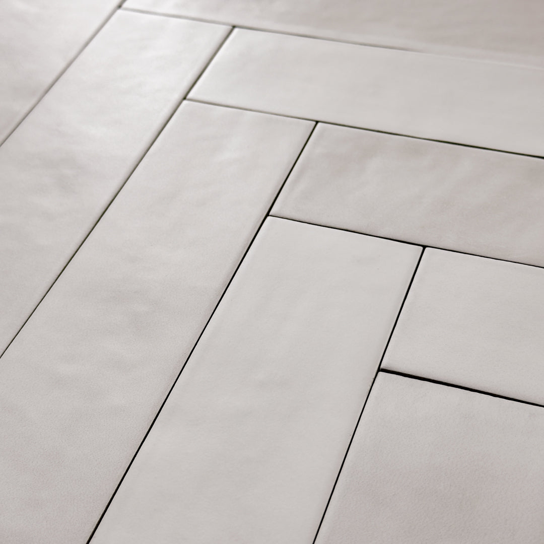 Angled close up view of 2x9 matte subway tiles in a light grey shade with subtle texture. Laid out in a herringbone pattern. 