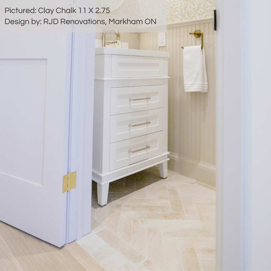 A look inside a quaint but luxury powder room, there is wallpaper on the upper and beige wainscotting on the lower walls. The floors are tiled in clay chalk brick stone look 11x2.75 tiles. they are laid in a herringbone pattern with two rows of brick around the perimeter. 