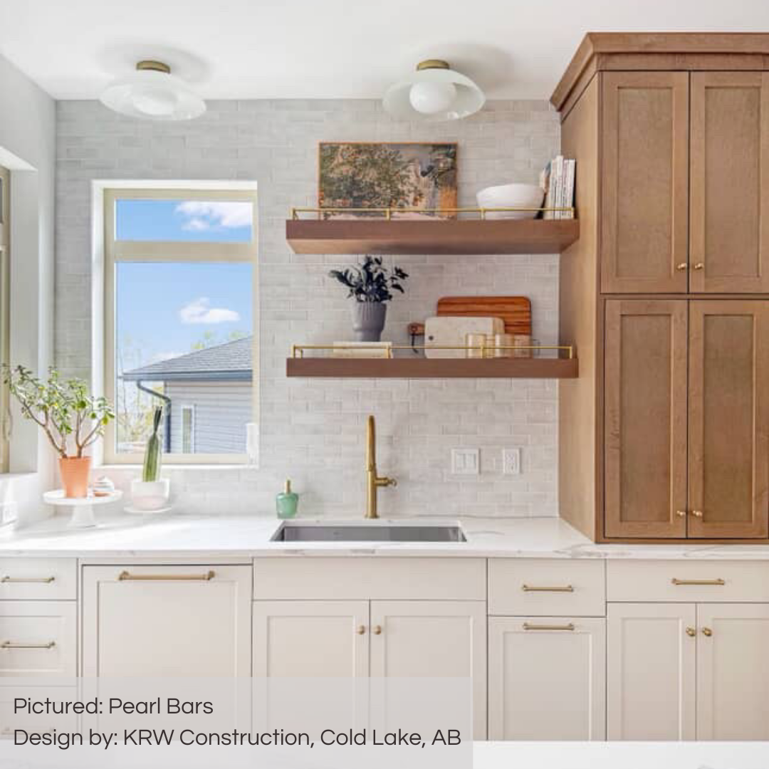 A delightful and bright kitchen with white fixtures and some wood shelving and closed cabinetry. the charming gold hardware adds elegance. from this perspective the room is filled with light from the two windows at the corner of the kitchen. the walls have been tiled with pearl bars variated off white subway tiles in a traditional brick layout. 