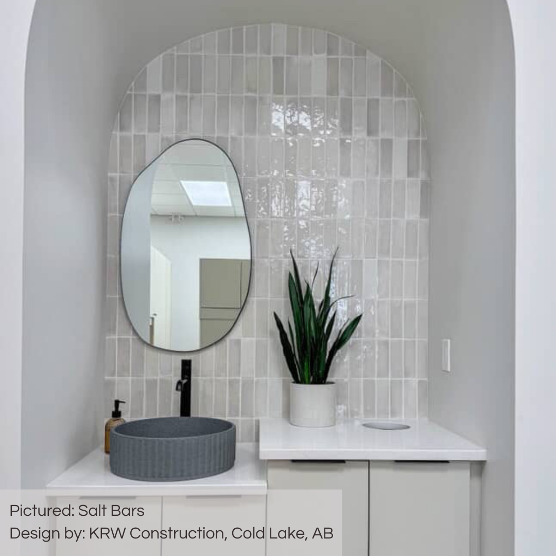 A dental office sink station with cabinets, sink and mirror within an arched nook. the back wall is tiled in salt bars glossy subway tiles, the tiles are heavily variated in shades of whites  greys and greiges. the tiles look cooler due to the shadows and surrounding materials like the blue stone sink! 