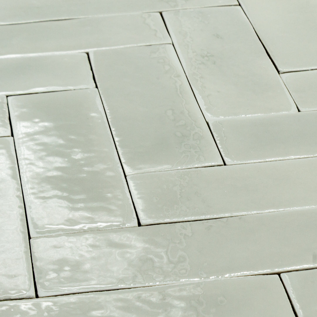 angled view of a group of 2x6 glossy light green sage subway tiles in a herringbone layout. the edges are slightly uneven to provide a subtle texture and light bounces off them