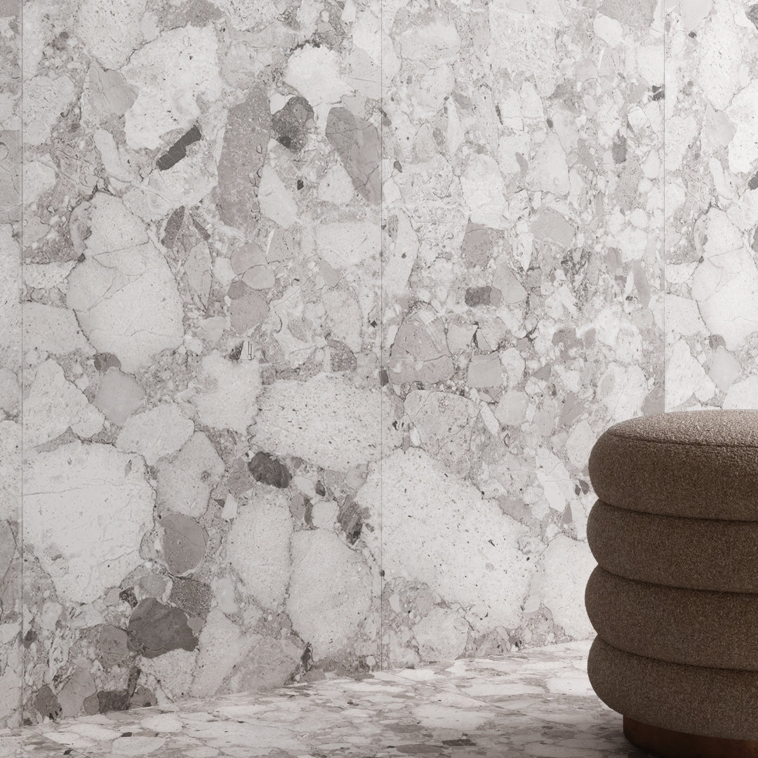A floor and wall application using the same tile for a natural look. The tile is 24x48 Rainstone mist light grey terrazzo look porcelain tiles. There is a beige fabric stool in the corner of the image. The warm grey and beige shades compliment eachother.