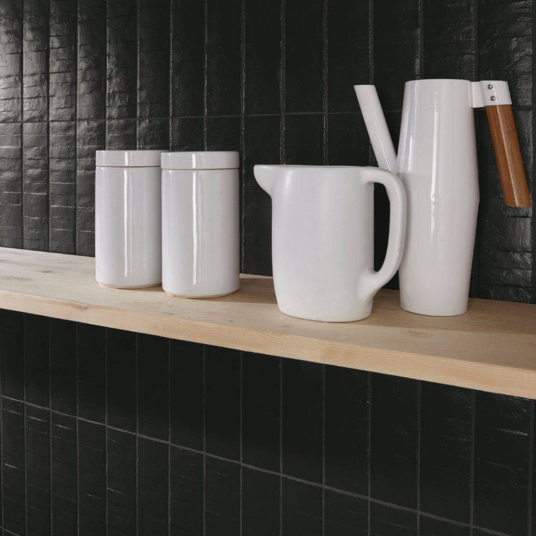 A modern cafe backsplash using black 3x12 textured tiles in a trendy vertical stack layout. The light wood shelf and white barware pop with contrast. 