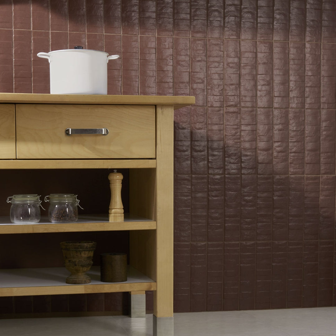 A close up of a tiled kitchen wall, the tiles are wine red burgundy with textured in a 3x12 size and the layout is a modern vertical stack. 