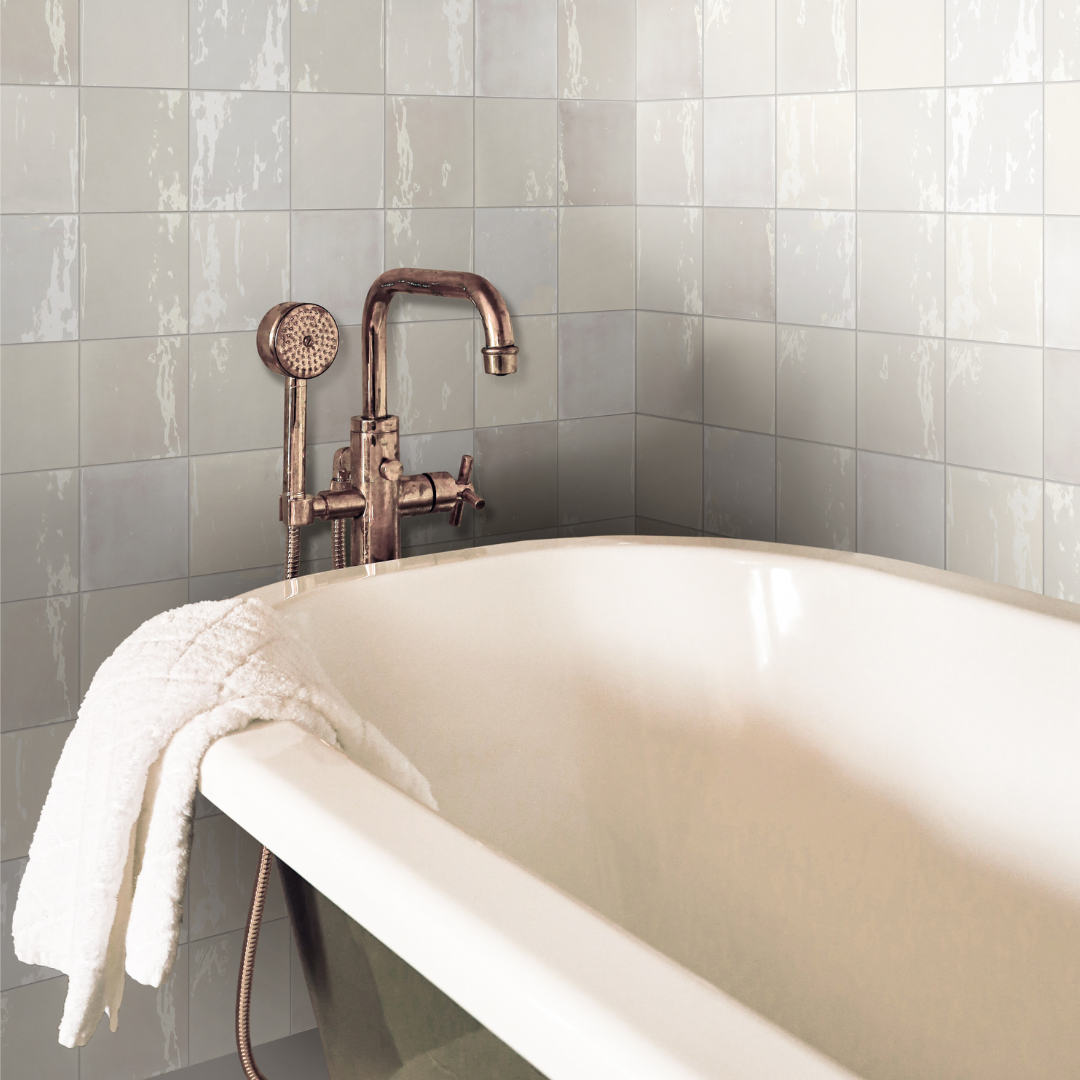 A close up of a clawfoot bathtub with tiled walls surrounding. The tils on the walls are light grey with colour variation, a glossy finish that catches the light and a subtle texture. 