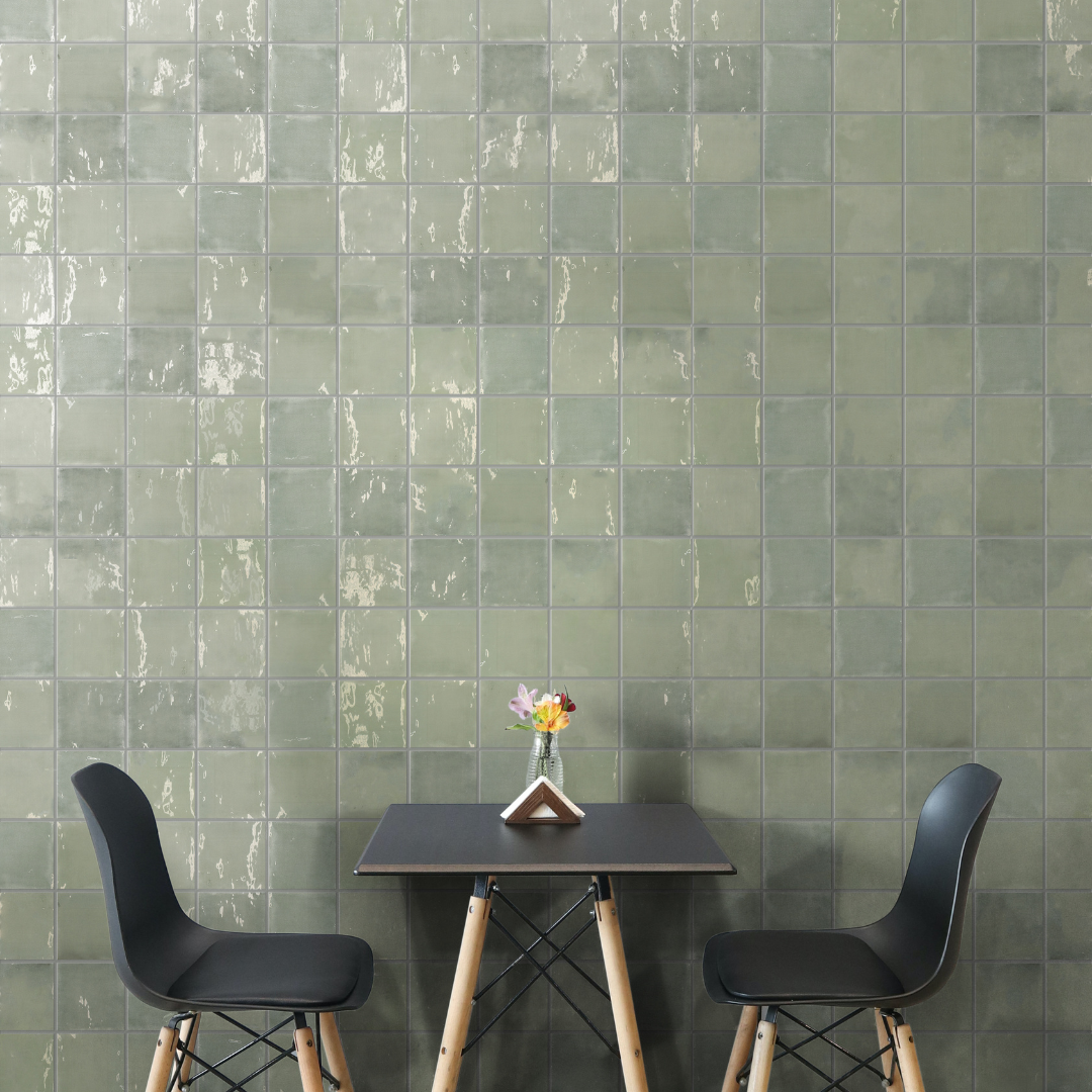 A table that seats two against a tiled wall. The tiles are a glossy 5x5 that have a subtle texture and catches the light. The colour of the tiles vary from shades of green in an earthy look. 