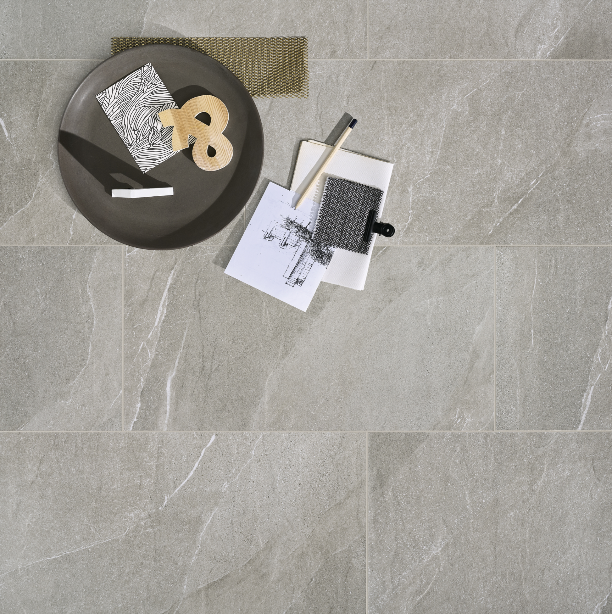 Ariel view of a large format rectangular limestone look tiles in taupe with light veining and a matte finish.  there are home accents and textile samples layered over the tiles. 