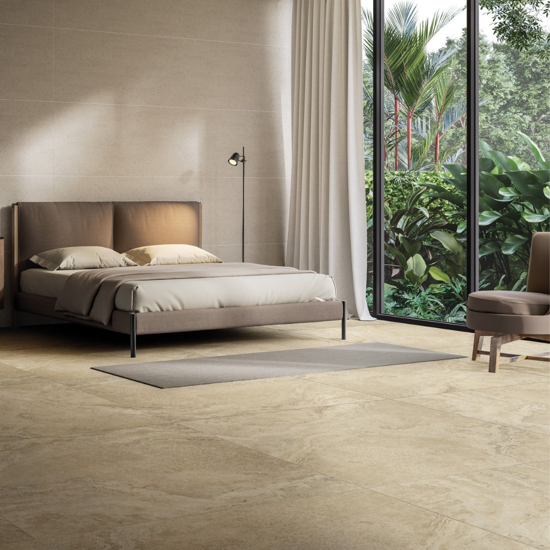A minimalist modern bedroom with neutral tones in the textiles and furniture. There is a large window with greenery on the other side of the glass. The tiles floor looks like a rich travertine stone in a warm beige shade but it is durable porcelain. 