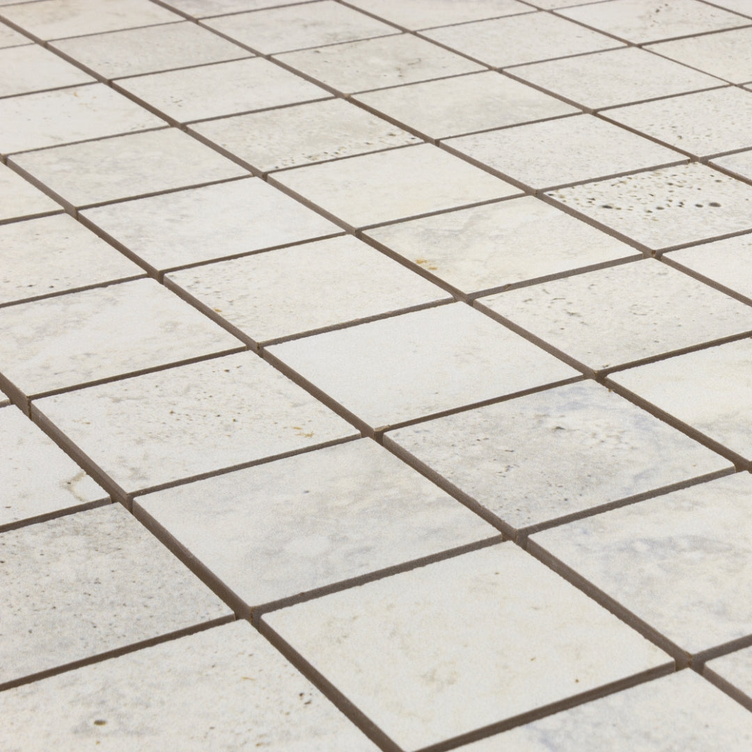 Angled close up of a 2x2 square mosaic tile that resembles travertine stone in light grey white. 
