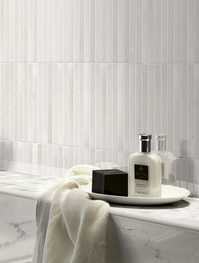 Close up on a bathroom shower wall featuring 5x10 panels of white glossy fluted tile. It is paired with marble for a luxurious look. 